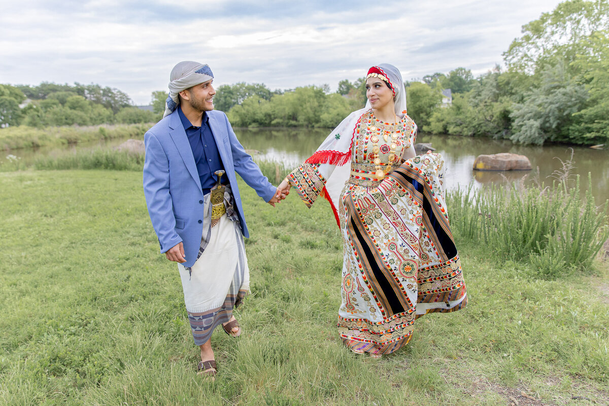 A couple holds hands as they walk through a grass field, behind them a river. They are wearing traditional Palestinian clothing.