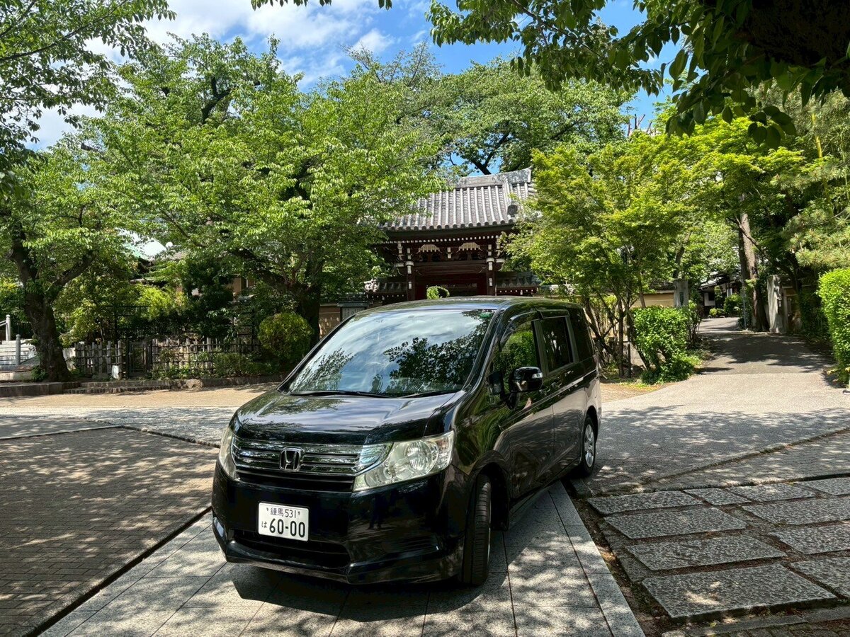 A black Honda Stepwgn parked in a serene Japanese temple courtyard surrounded by lush greenery and traditional architecture under a sunny sky.