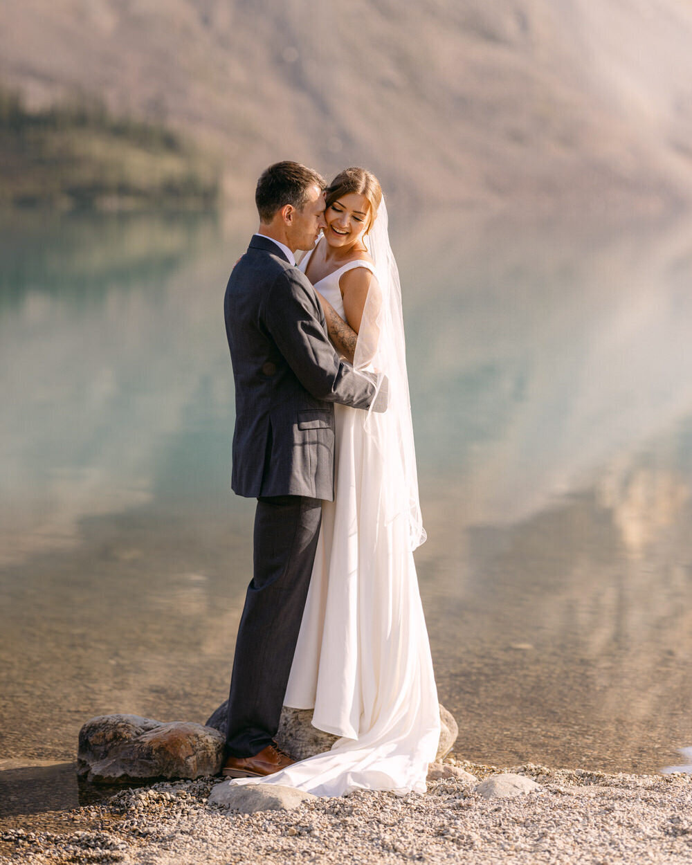 This stunning photograph captures the bride and groom standing on a rock at Bow Lake in Banff National Park during a beautiful summer evening. The still waters of the lake and the soft glow of the evening sun create a serene and romantic atmosphere, enhancing the beauty of the moment.
