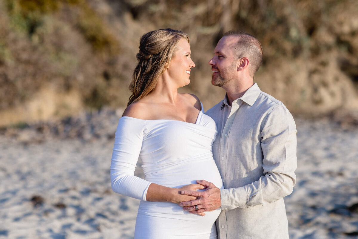 A pregnant woman in a white dress stands on a sandy beach with a man in a light shirt. They are gazing at each other, smiling, as he gently places his hand on her baby bump. The background is a blurred mix of sand and greenery.