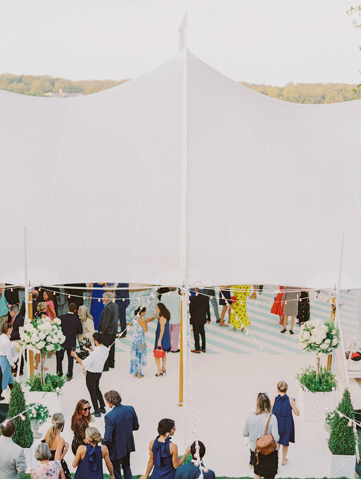 The photo was taken at The Hamptons, New York, by photographer Bonnie Sen. A crowd of people gathers under a white event tent decorated with hanging lights and floral arrangements at the beautiful waterfront estate. Individuals are dressed in semi-formal attire, with some wearing lovely blue dresses that complement the sunny outdoor setting perfectly. Videographers from East West Films were also present to capture the moment.