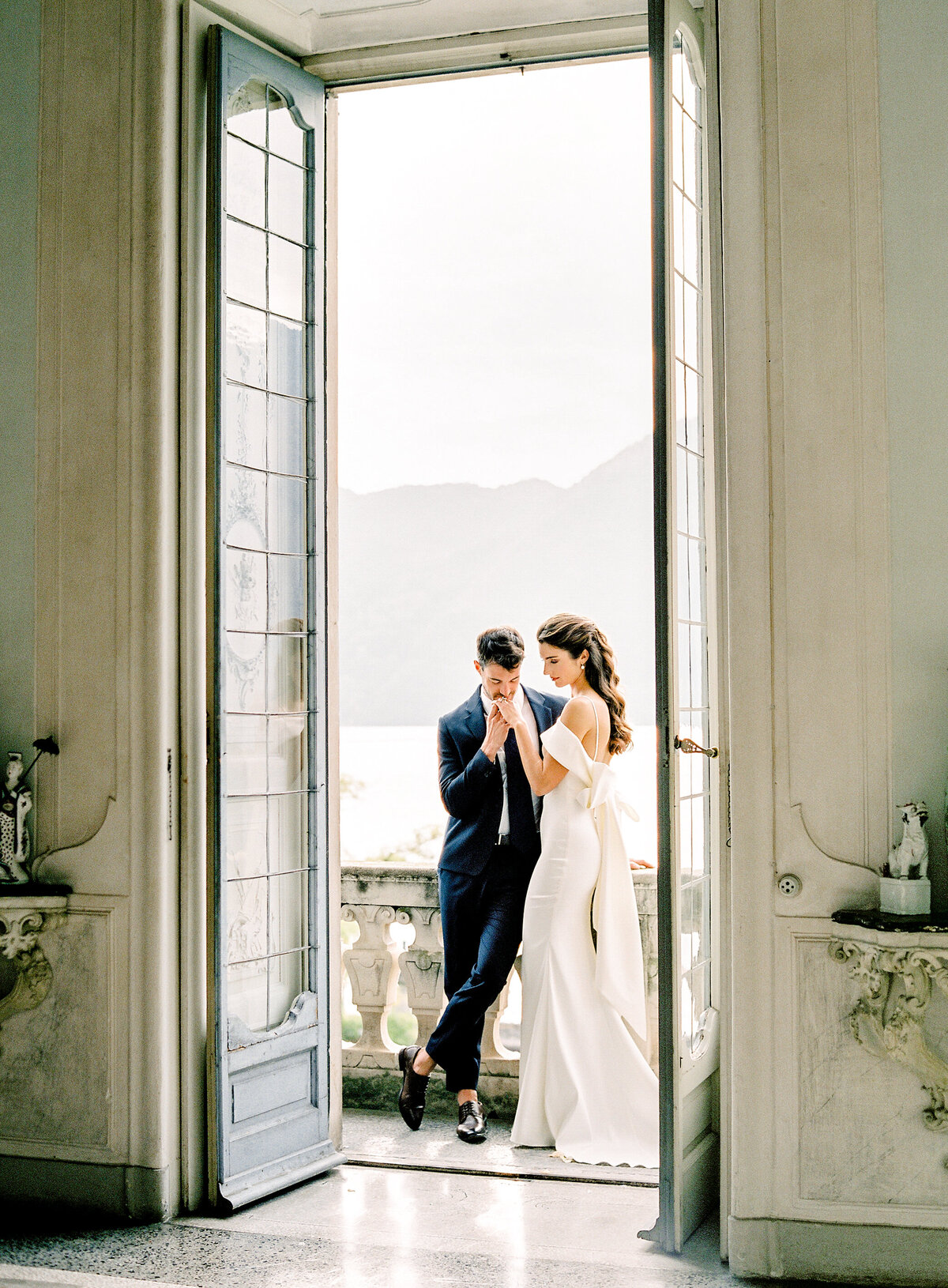 Bride and groom in an intimate moment on the balcony of Villa Sola Cabiati on Lake Como bride in an off the shoulder wedding dress with oversized bow in the back and groom in navy suit during their Lake Como wedding photographed by Italy wedding photographer