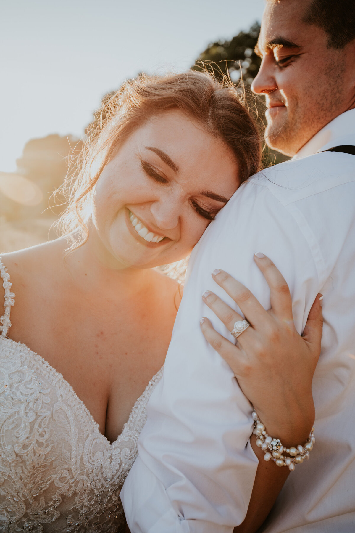 Bride leans on grooms shoulder while holding his arm as the sun rays shine.