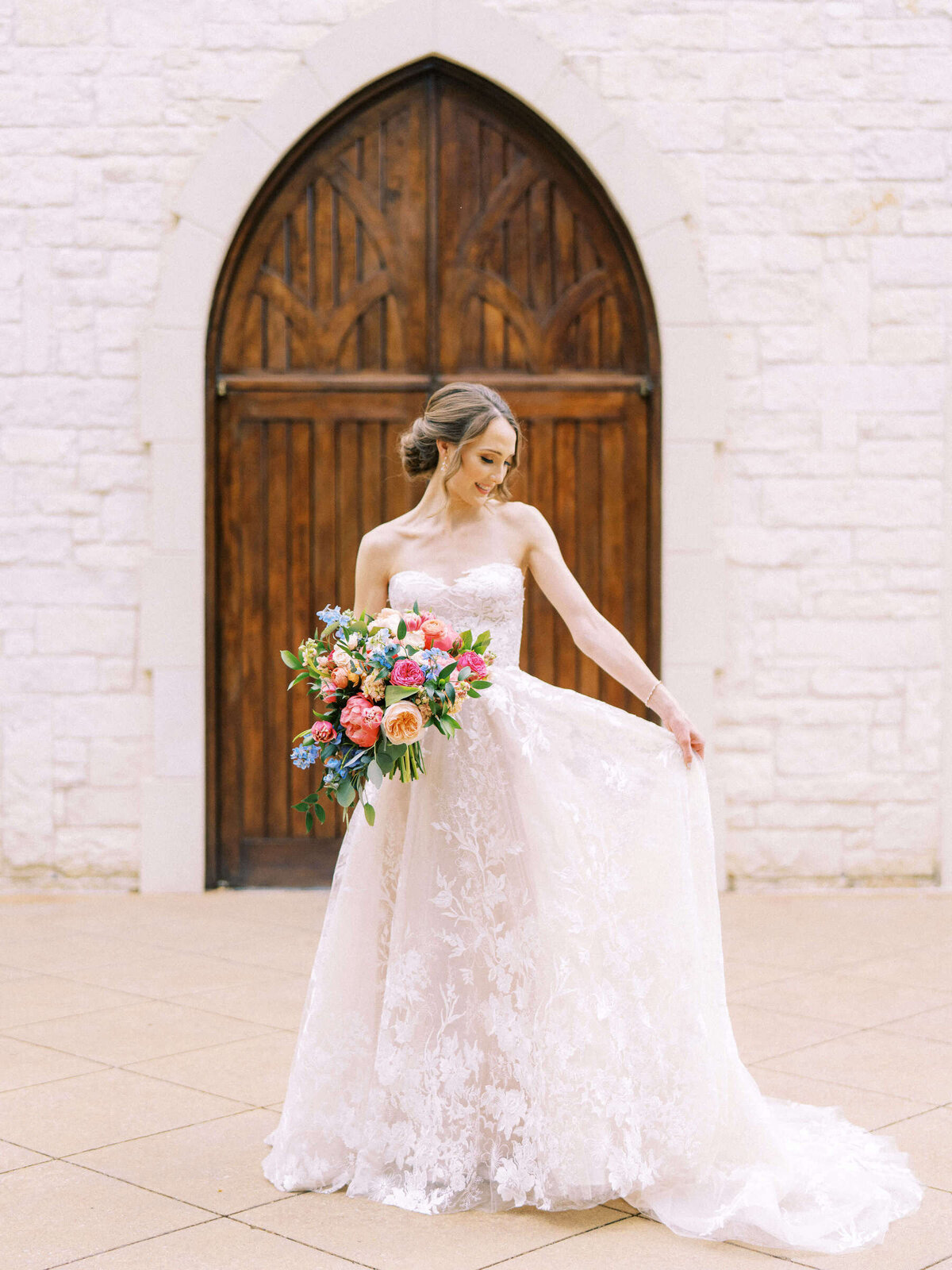 Bride stands in front of arched wedding chapel doors in Monique Lhuillier wedding dress holding colorful bouquet