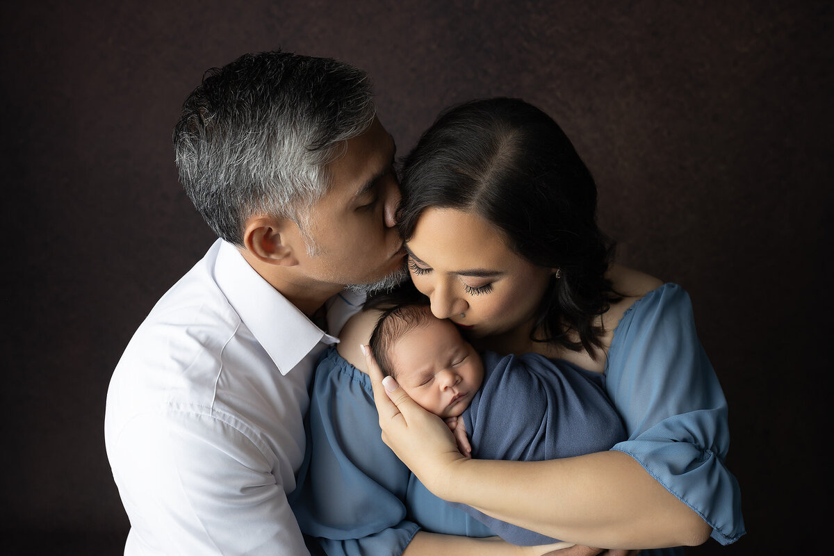 Mother and father kiss their newborn while wrapped in soft blue, capturing the tenderness of early parenthood.