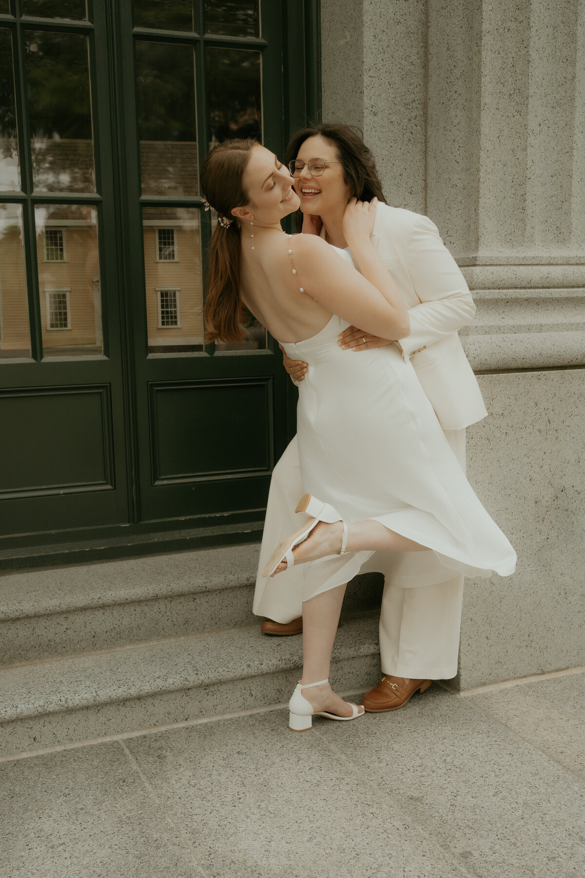 Joyful wedding same sex couple standing on stone steps, embracing in front of a green door. The bride, wearing a sleeveless white dress with a pearl hairpiece, leans back while the other bride, dressed in a stylish white suit, holds her close, both sharing a happy moment.