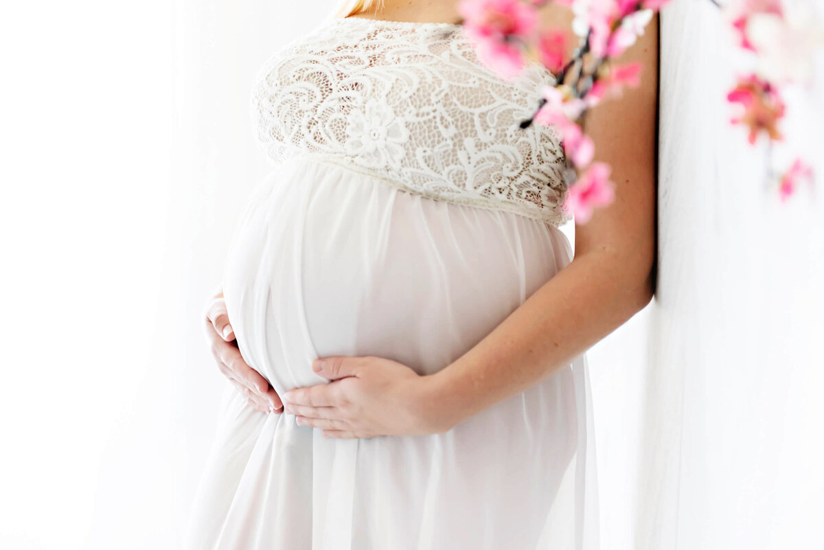 pregnant woman wearing a white maternity dress holding her belly for her in-studio maternity photos in Burlington