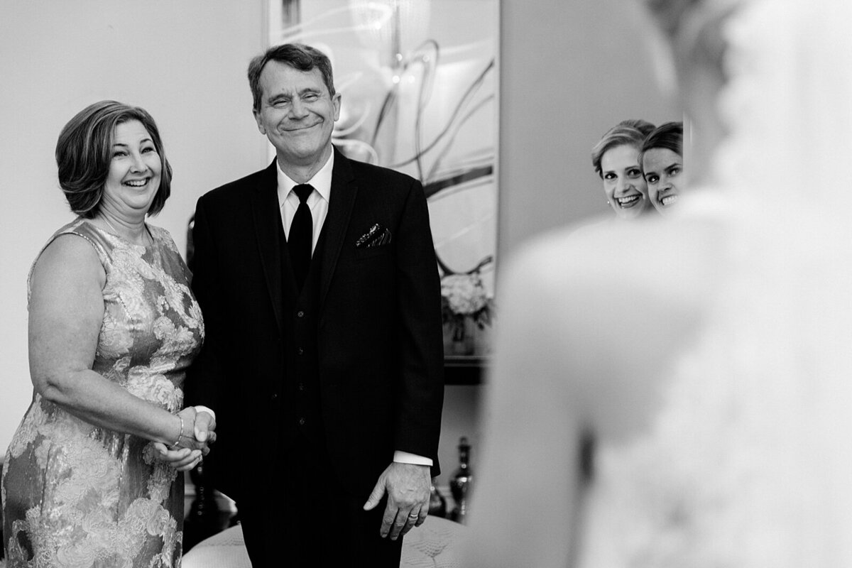 Candid first look with the bride and her father, joined by her mother, while two bridesmaids peek from behind a wall, capturing their joyful reactions