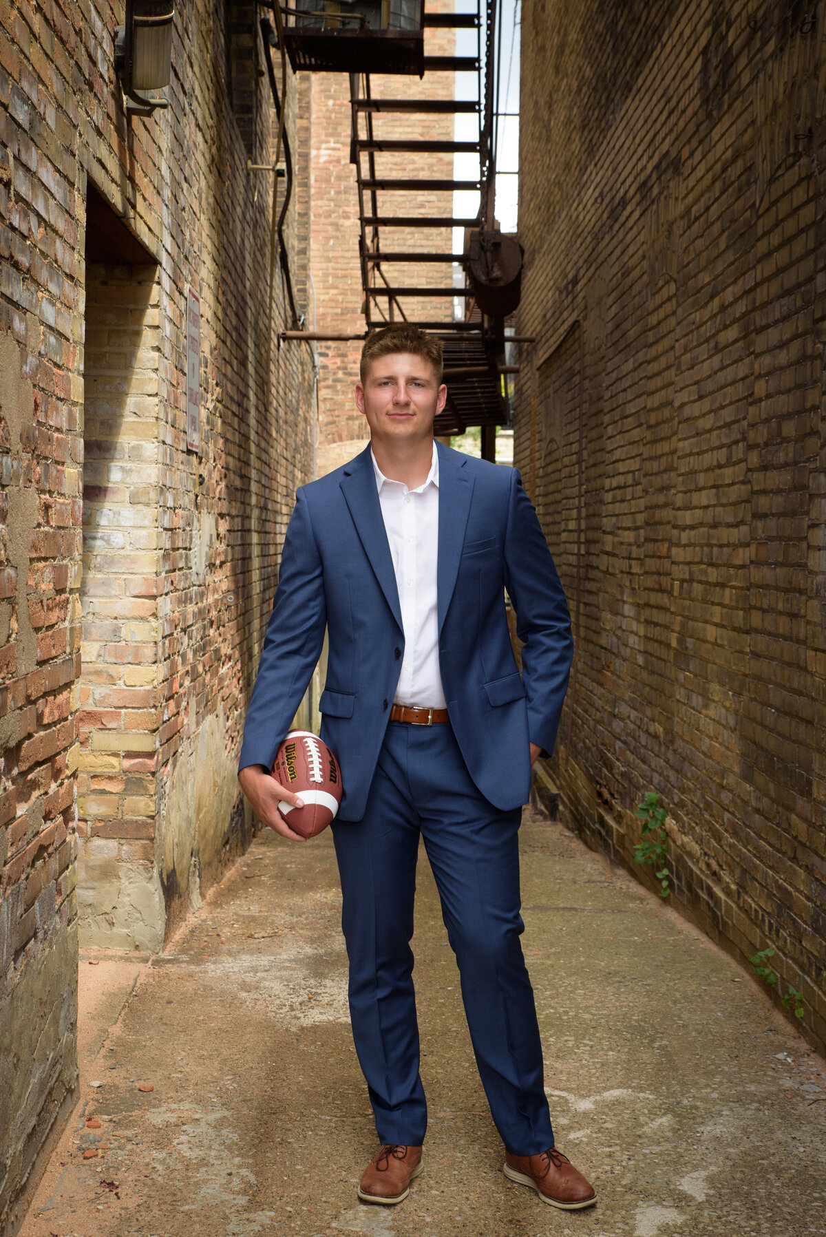 De Pere High School senior boy wearing a blue suit with white shirt holding a football in a brick alley in an urban setting in Downtown Green Bay, Wisconsin