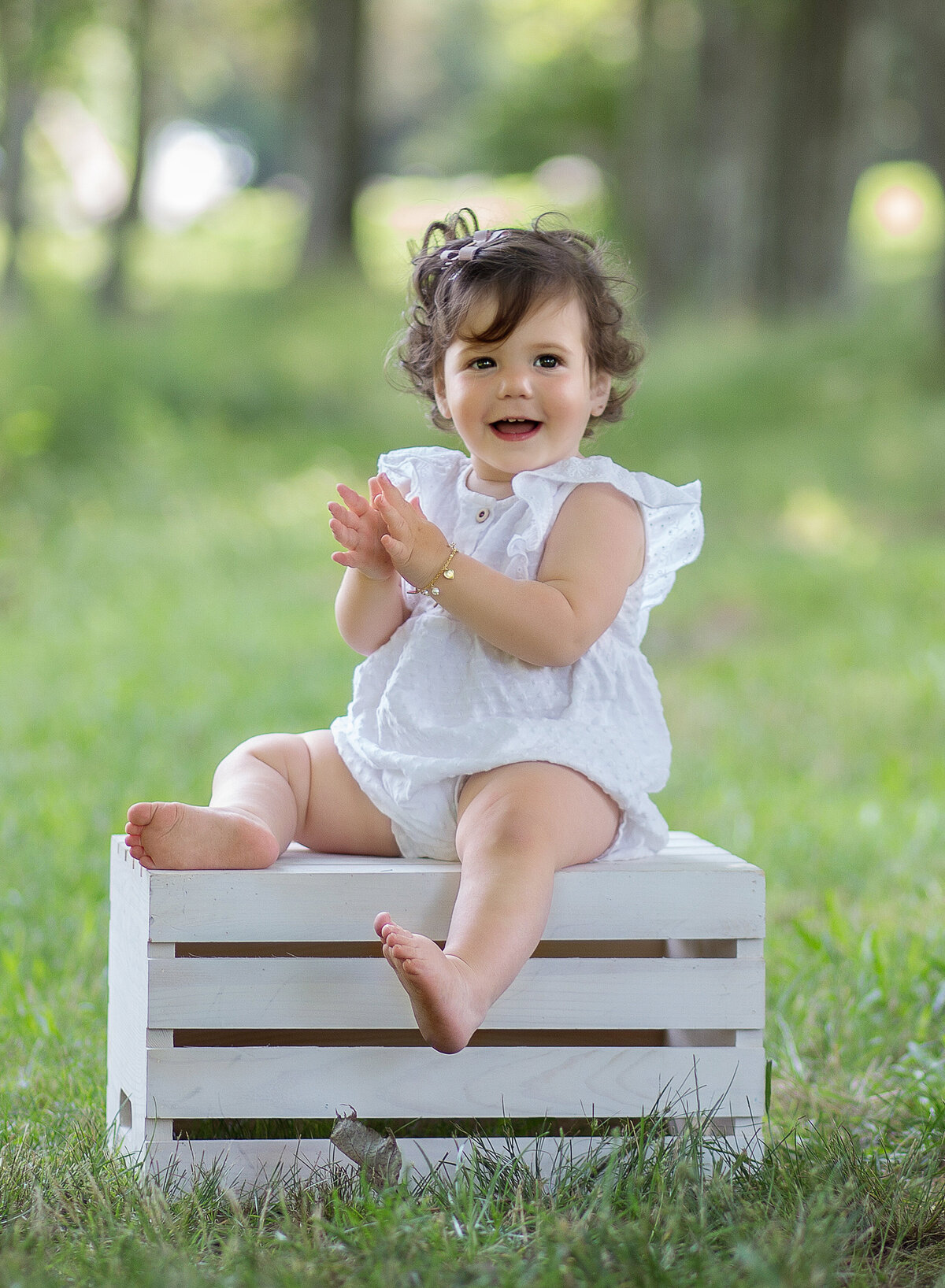 Little girl sits on a white milk crate in a brooklyn park for a family photoshoot. She is looking at the camera and clapping. Captured by Chaya Bornstein, Brooklyn Family Photographer