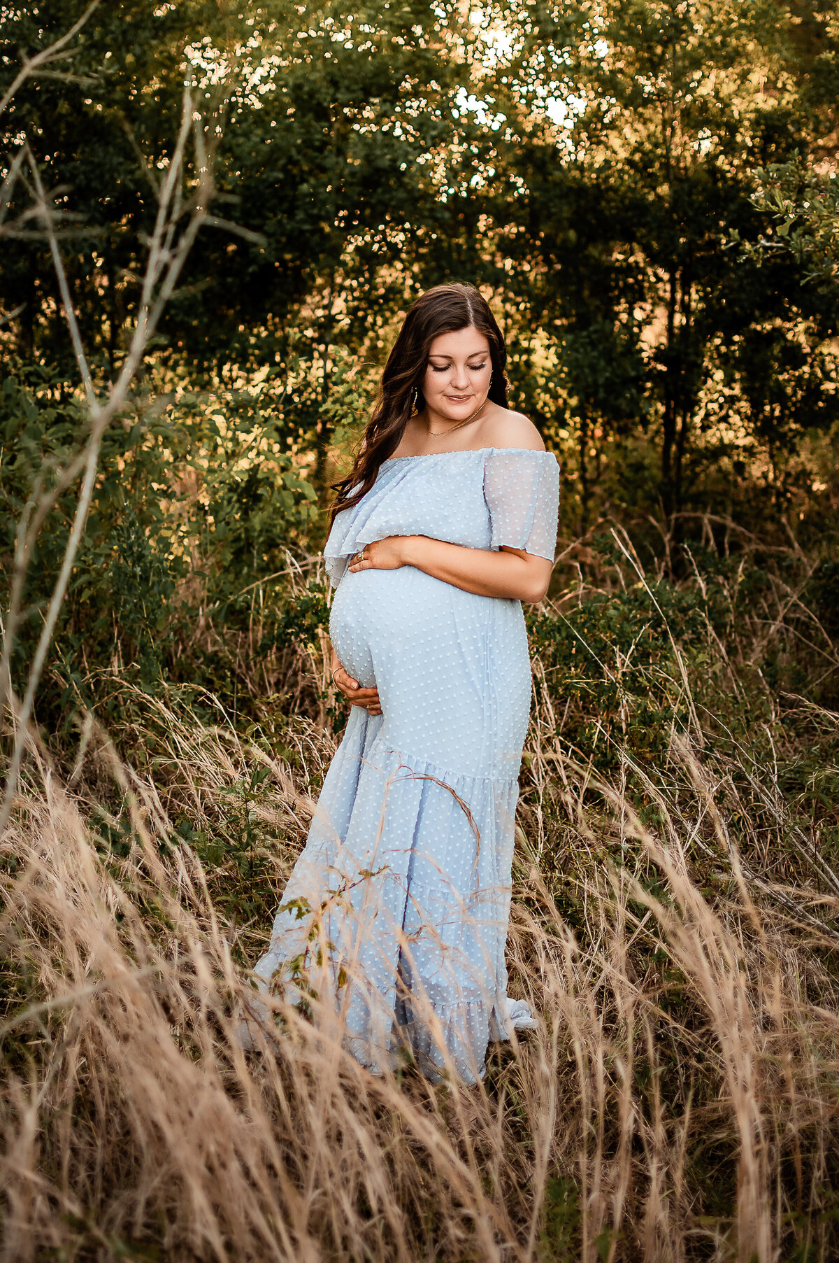 An expectant mother holds her belly and smiles down at her shoulder as she stands in a field of golden grass.