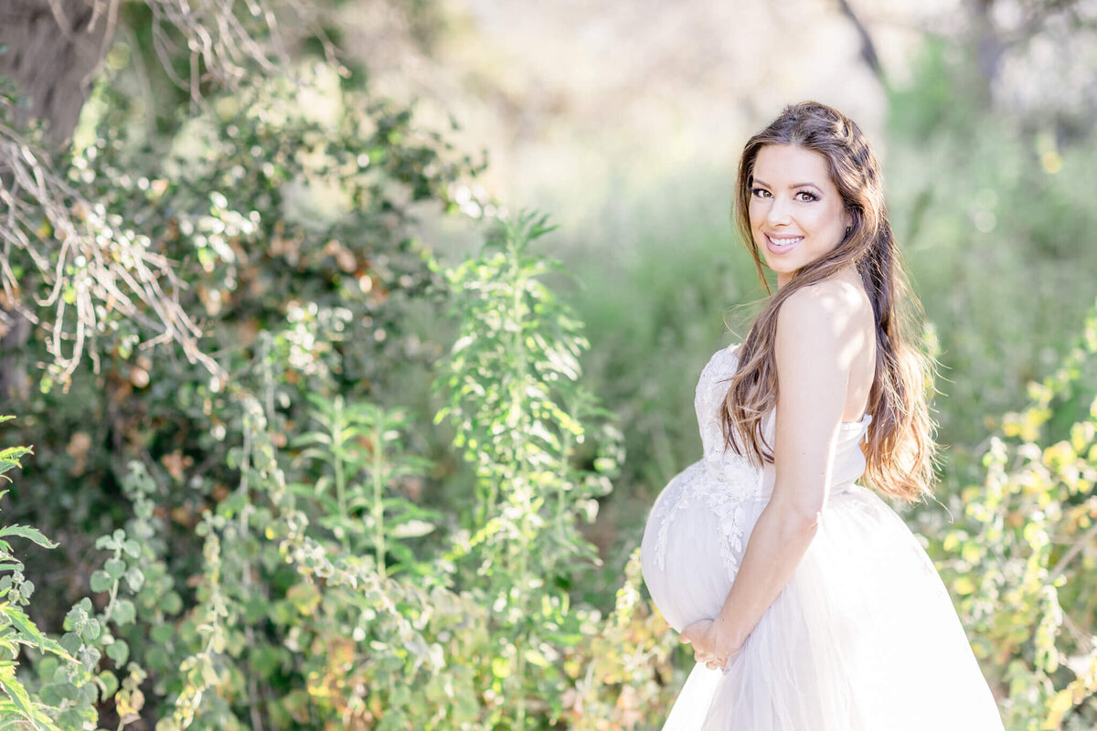 A pregnant woman in a flowing white dress stands in a lush, green outdoor setting, smiling and looking over her shoulder. Sunlight filters through the trees, creating a soft, serene atmosphere.