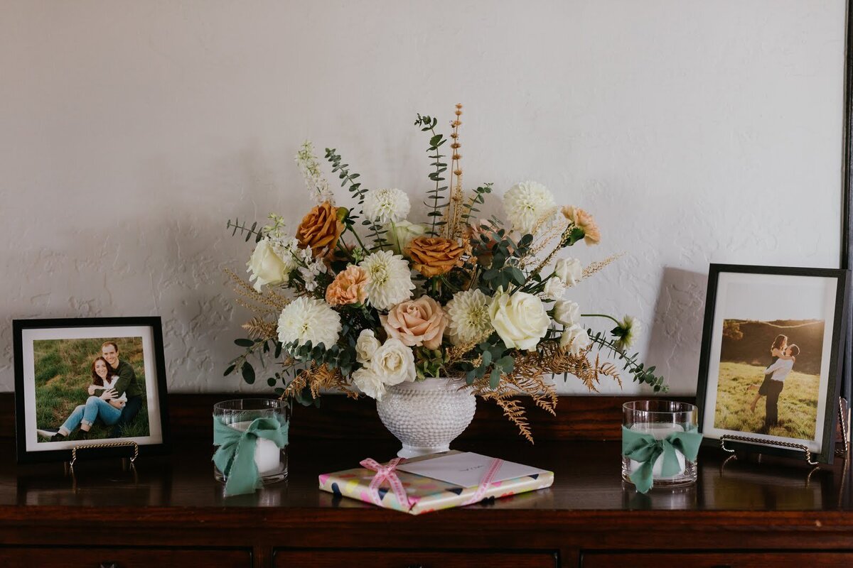 Mid sized floral arrangement sits atop a table with family photos at a wedding reception.