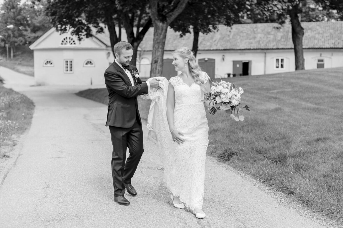 Brudgum håller upp brudens klänning när de går