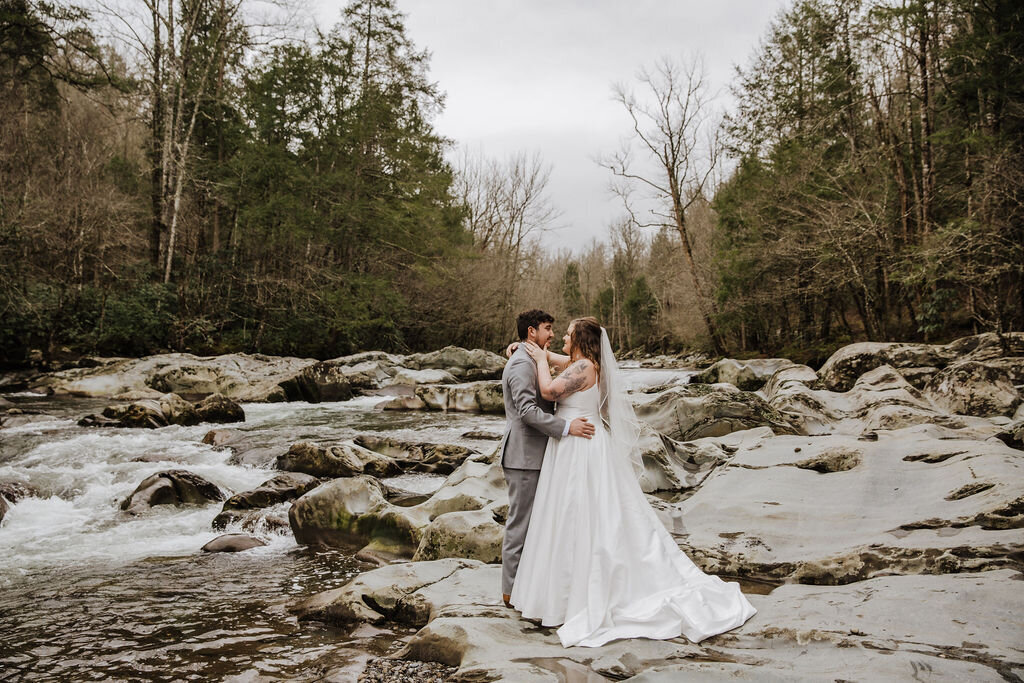 Bride and groom standing together on a rocky riverbank at Greenbrier during their eloping to Gatlinburg, surrounded by rushing water and evergreen trees under a cloudy sky.
