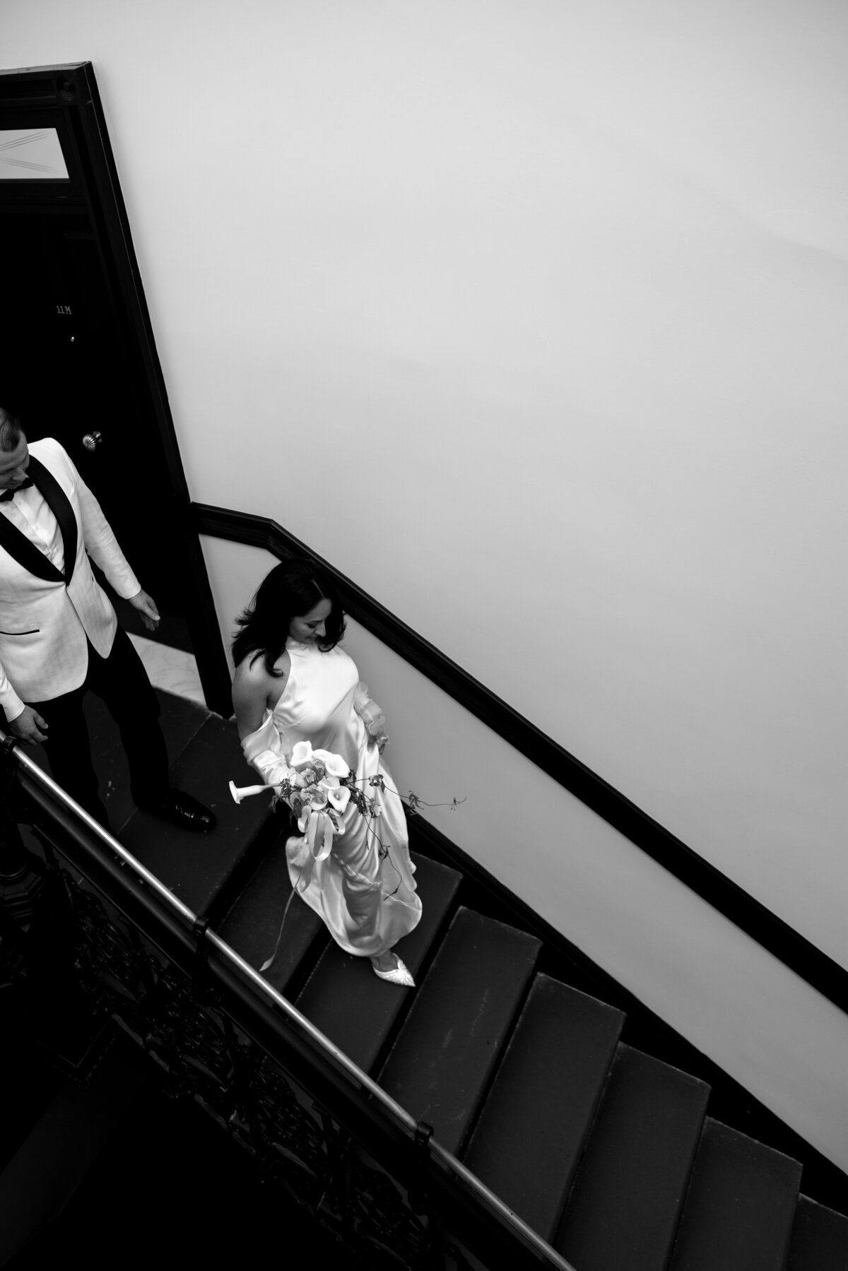 Bride walks down a modern staircase with sleek black railings while groom waits at the bottom during their NYC elopement.