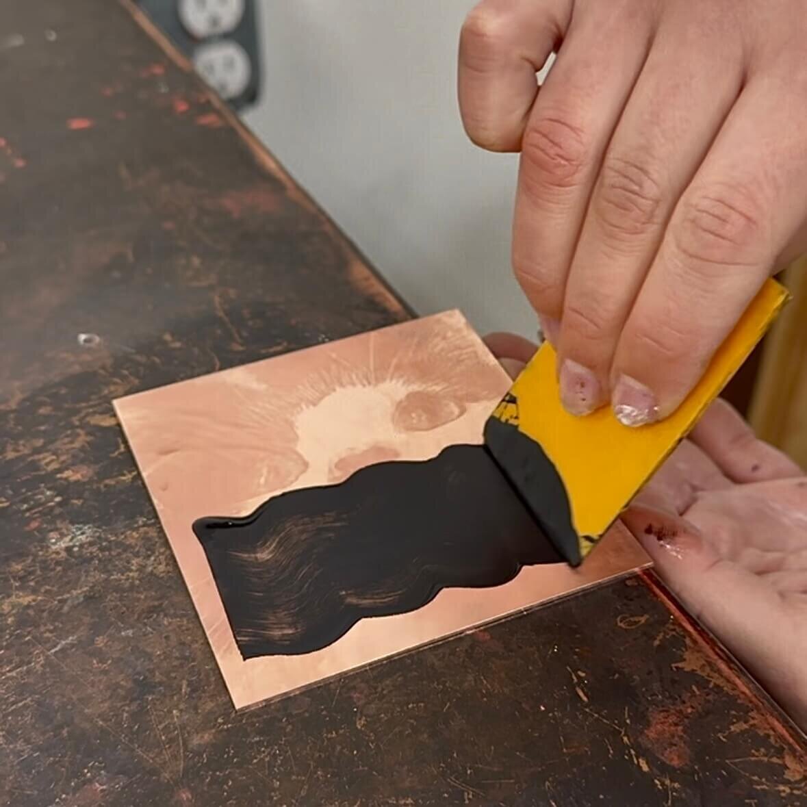 Close-up of a hand inking a copper plate etched with a cat image using black ink and a yellow squeegee.