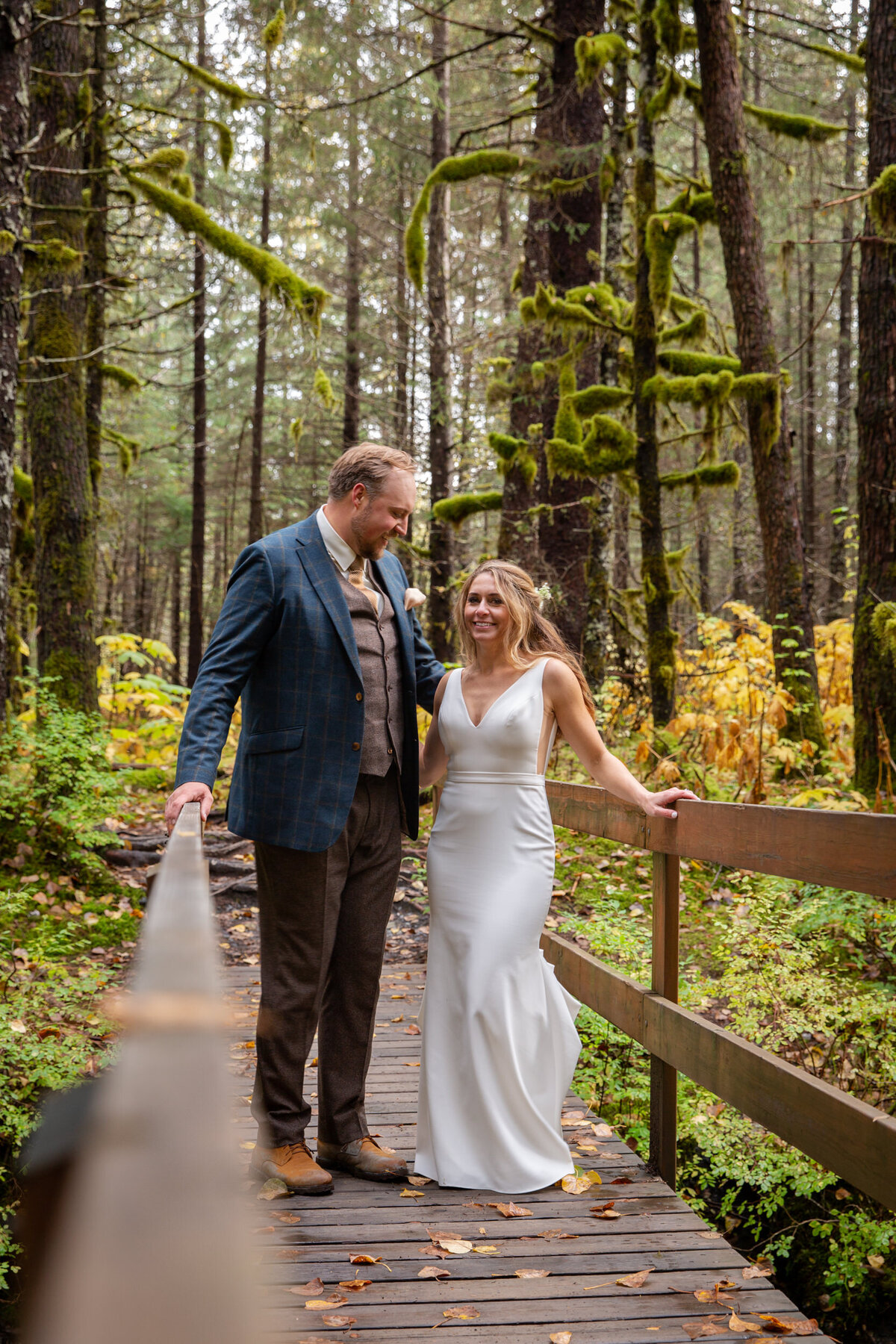 A bride and groom walk across a bridge in a forest in alaska.