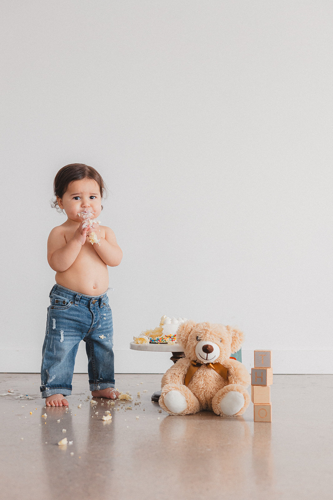 A little boy stands in jeans next to cake.
