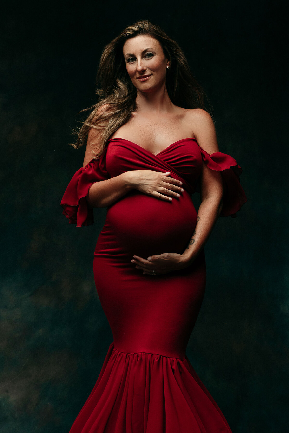 Maternity portrait of woman with long dirty blonde hair wearing maroon dress holding baby bump while smiling and standing on dark gray background