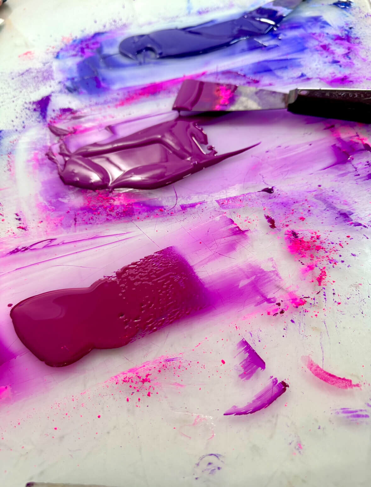 Smooth purple and magenta inks being worked and blended on a mixing surface, surrounded by bursts of pigment dust and color swatches.