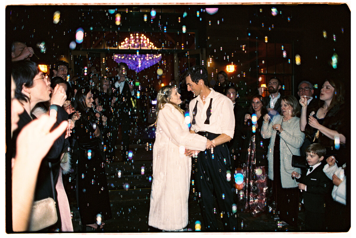 Bride and groom exit their wedding celebration surrounded by bubbles, captured on film in a joyful moment