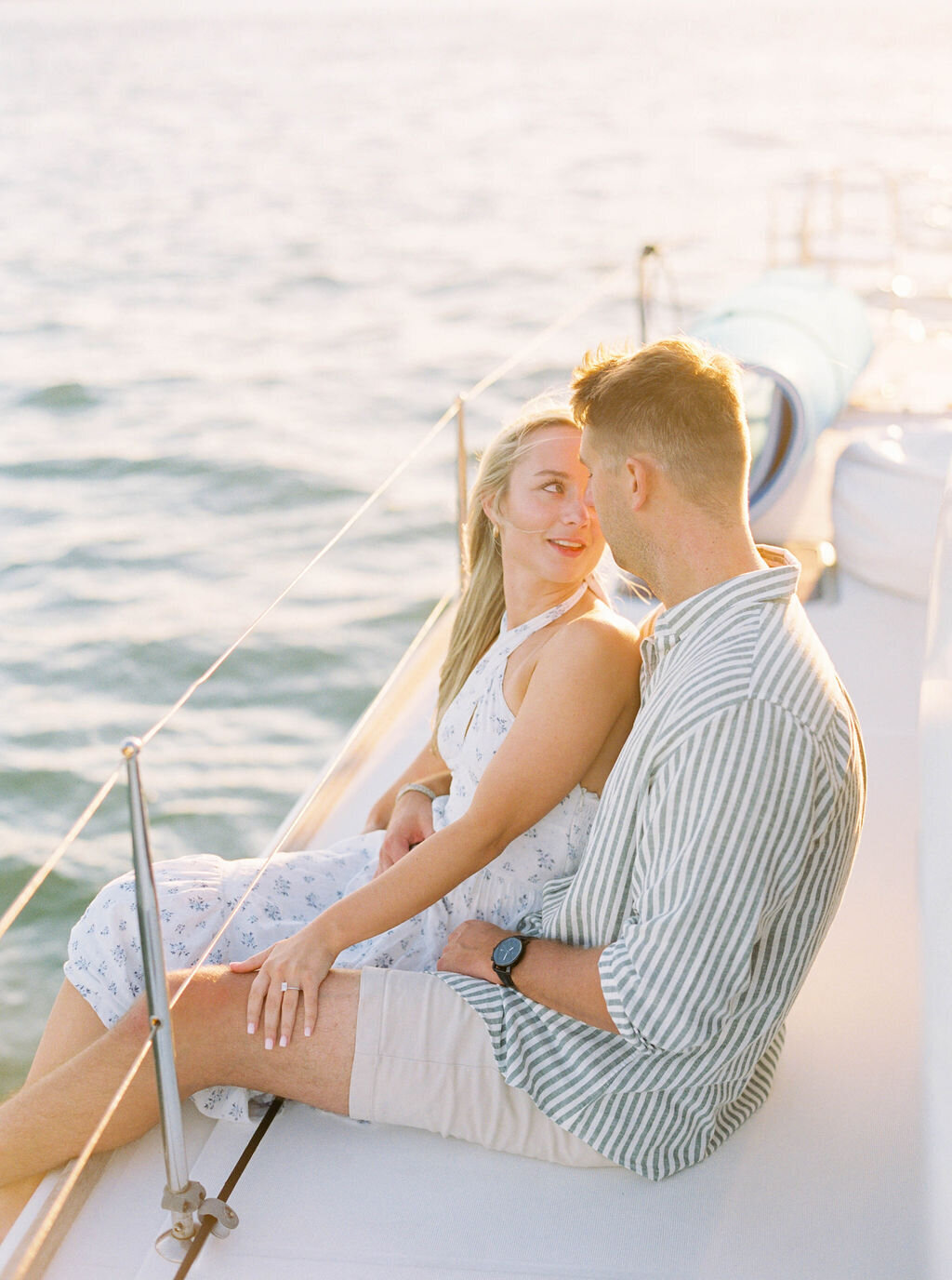 Newport Rhode Island Engagement Photo Photographer | A couple sits on a sailboat deck, gazing at each other lovingly. The sun casts a warm glow over the calm water