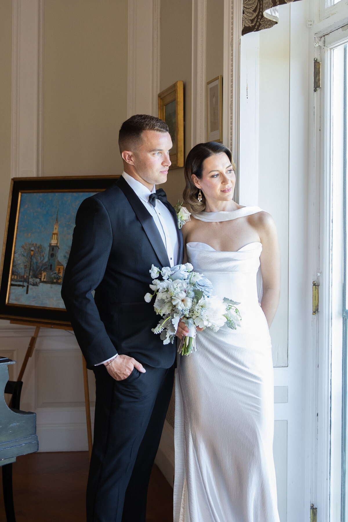 Aldrich Mansion Wedding Photos | Kelsey Sheehan Photography Timeless Rhode Island Weddings | A couple in elegant attire stands by a window. The woman holds a bouquet and wears a strapless white dress. The man is in a black tuxedo. A painting is behind them.