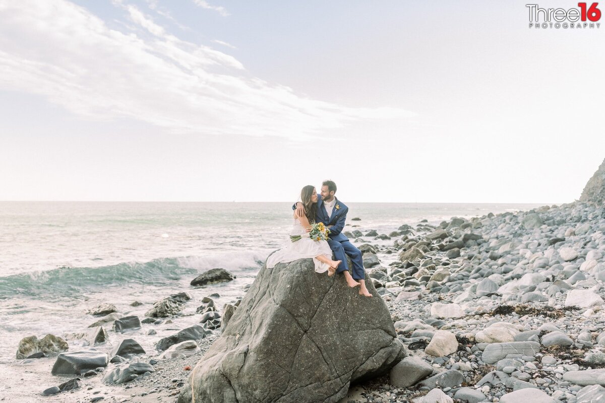 Bride and Groom go barefoot while sitting on a large rock together at the beach