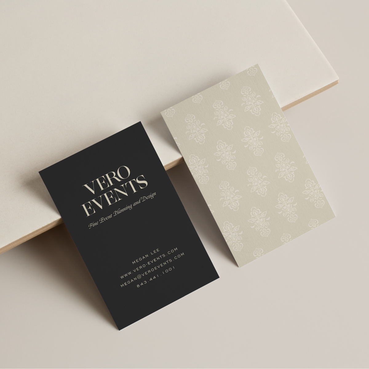 Two business cards, one black with white lettering and one beige with floral design, displayed against white countertop.