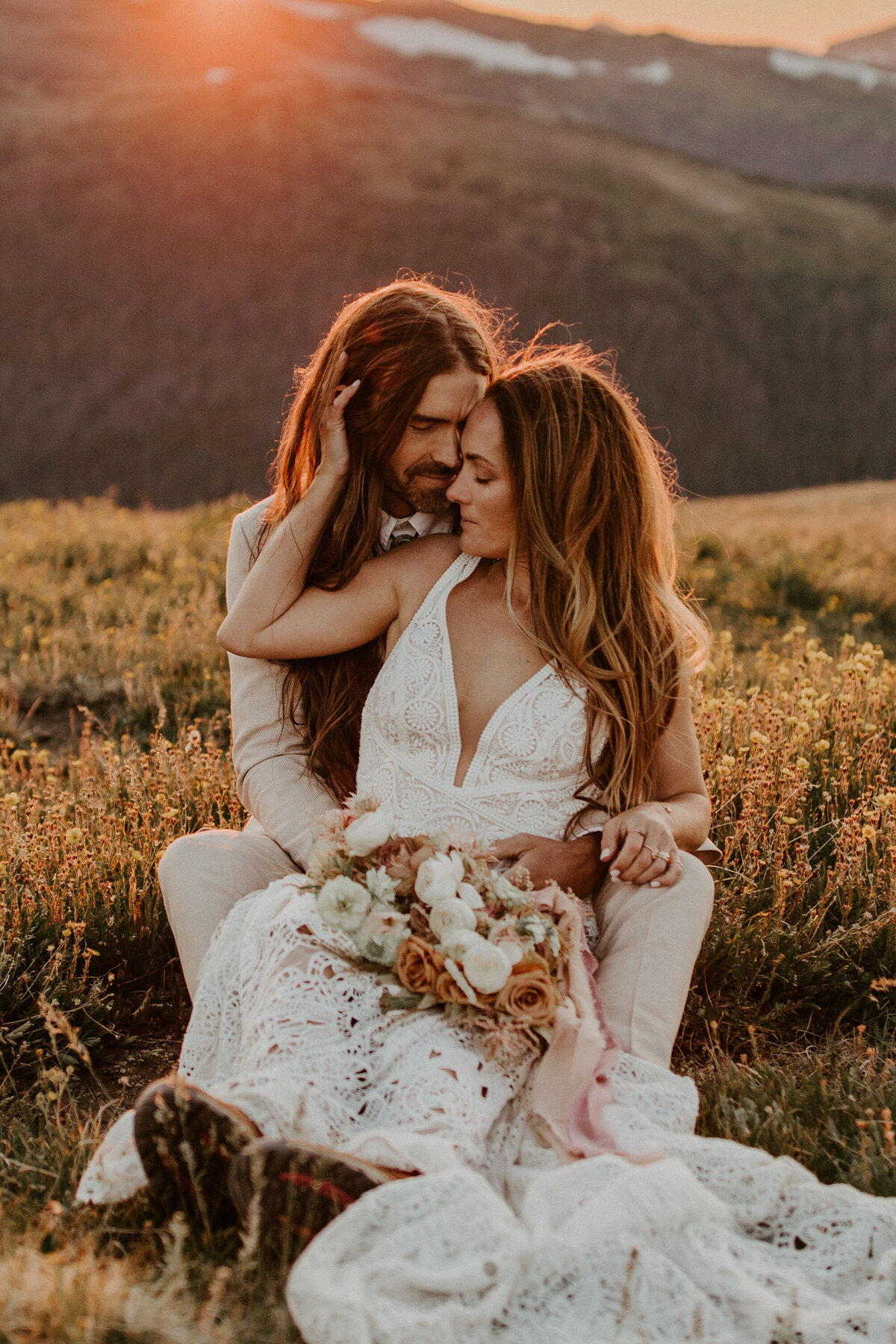 bride and groom pose holding each other with a bouquet while sitting on a field in the mountains