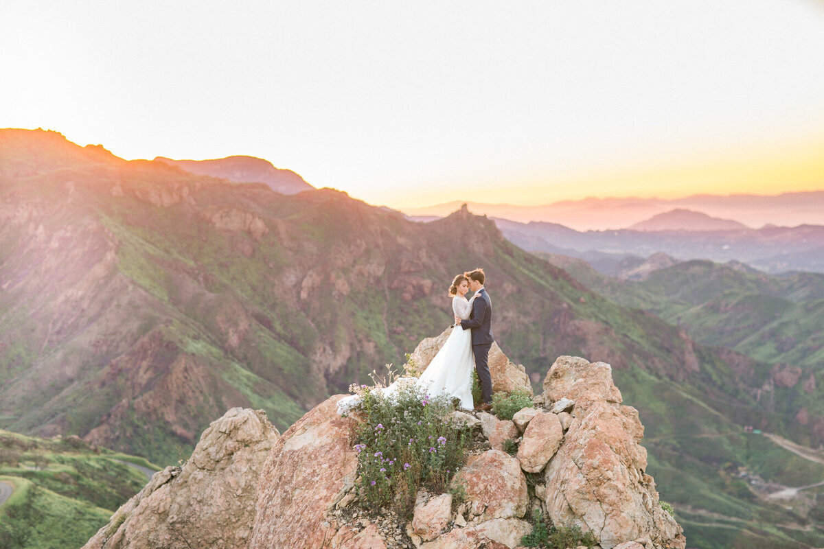 Kohnur couple on a rocky mountain peak at sunset. Bride in a flowy gown with a natural makeup look overlooking the hills in Malibu, California.