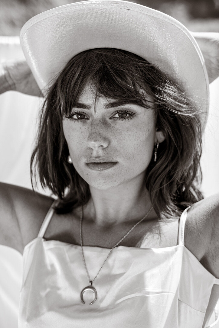 black and white portrait of a woman wearing a silver moon necklace and white cowboy hat with a satin silk cream top. She has brown hair and light complected skin with freckles