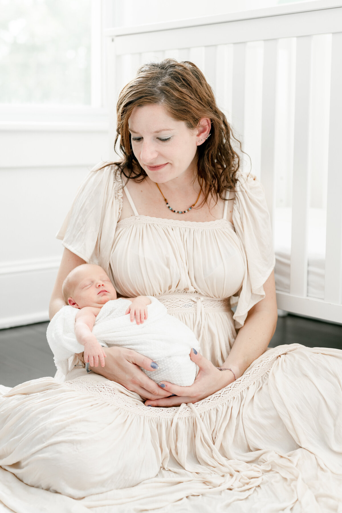 Mother sitting on the floor in a cream coloured dress with here newborn baby in her arm. Captured during her lifestyle newborn session in New Jersey