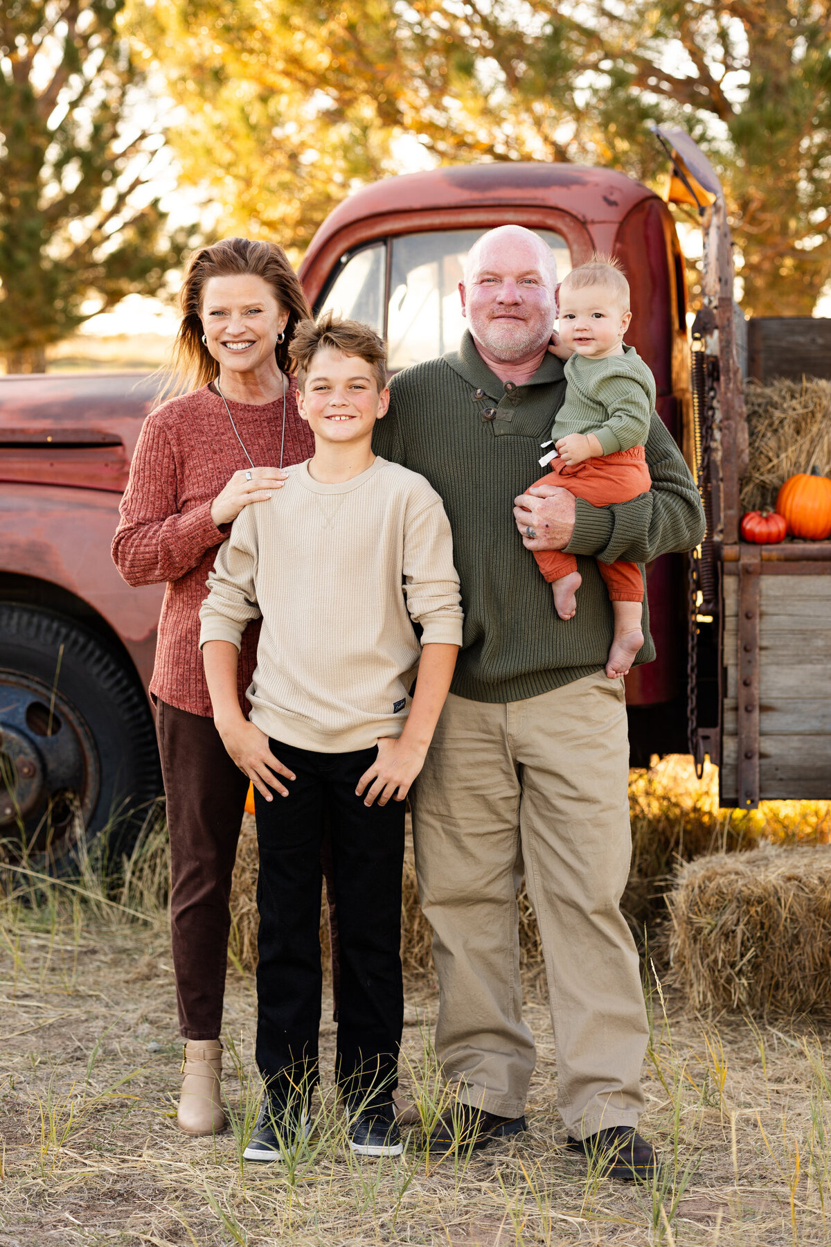 A grandma and grandpa stand with their pre-teen grandson and baby grandson in front of a vintage red farm truck and smile at the camera.