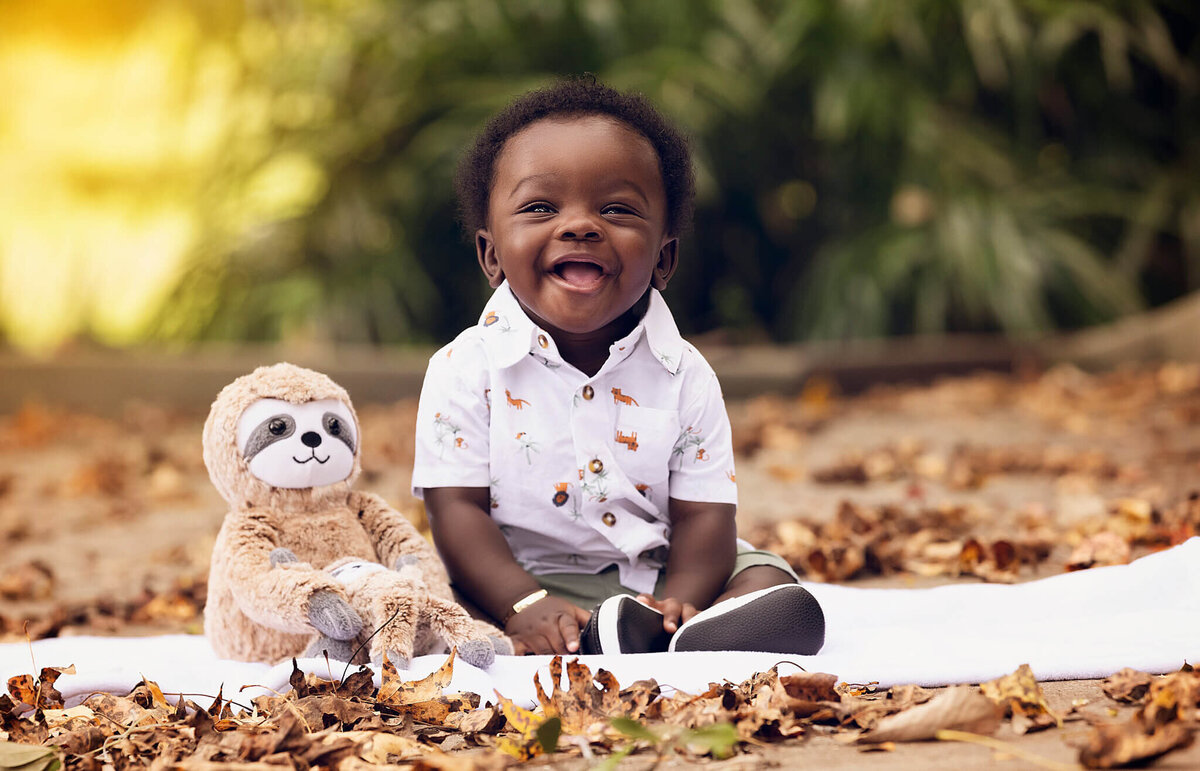 A joyful baby, captured during a milestone session in Orlando, wears a white shirt with animal prints and sits on a blanket outdoors. The baby smiles with an open mouth beside a plush toy sloth, surrounded by scattered autumn leaves and green foliage in the background.