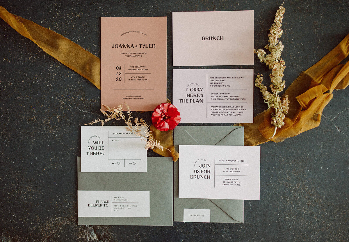 Mixed off-white, peach and mauve-colored wedding stationery with black font atop ribbon on concrete with wedding bands.