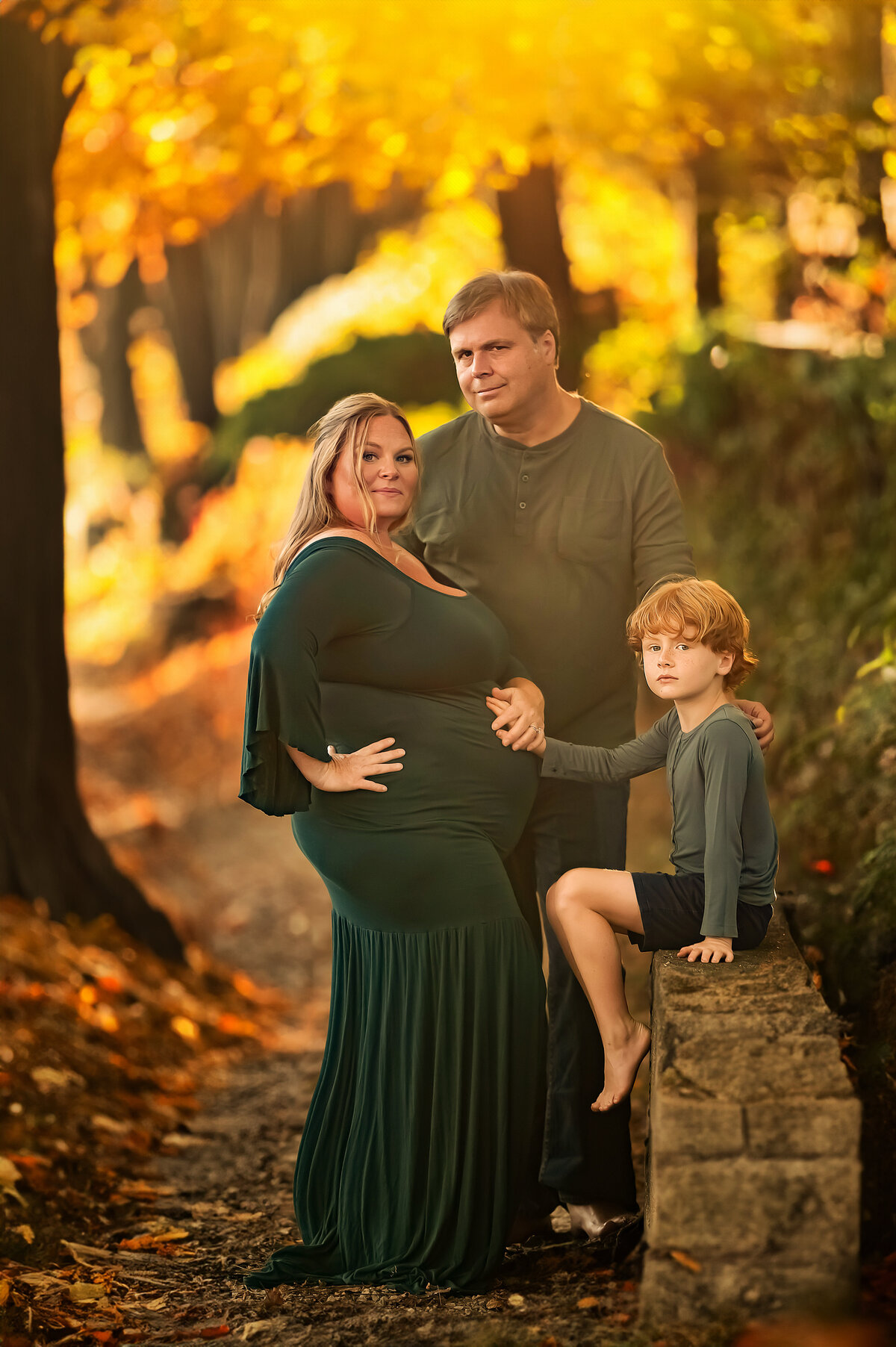 Pregnant mother in a green gown posing with her partner and young son in a golden forest path, surrounded by vibrant fall foliage during a family maternity session in Waukesha.