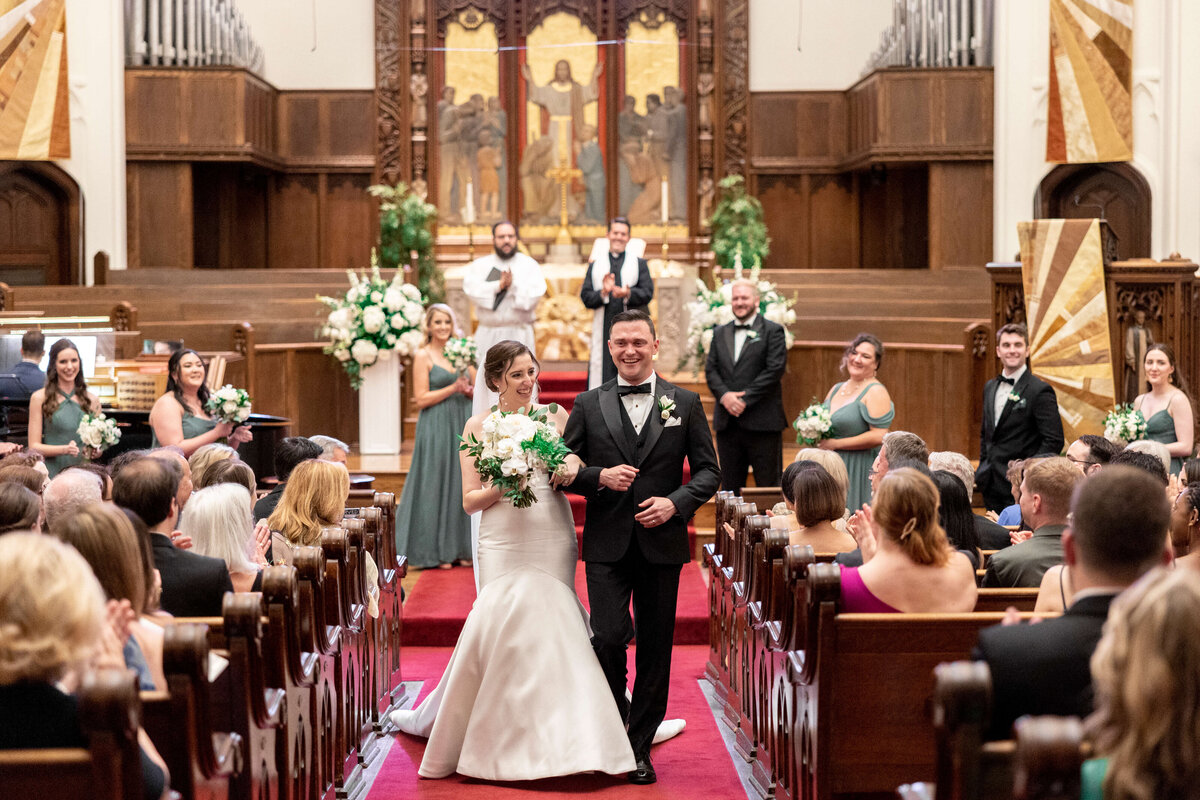 A bride and groom walk down the aisle of a church, smiling and holding hands. The bride carries a bouquet, and bridesmaids and groomsmen are in the background, clapping. The setting is ornate with wooden pews and a decorated altar.