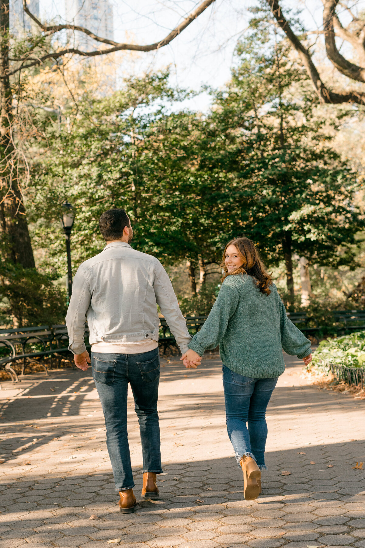 man and woman holding hands in park while walking away together and woman is looking back  smiling over her shoulder