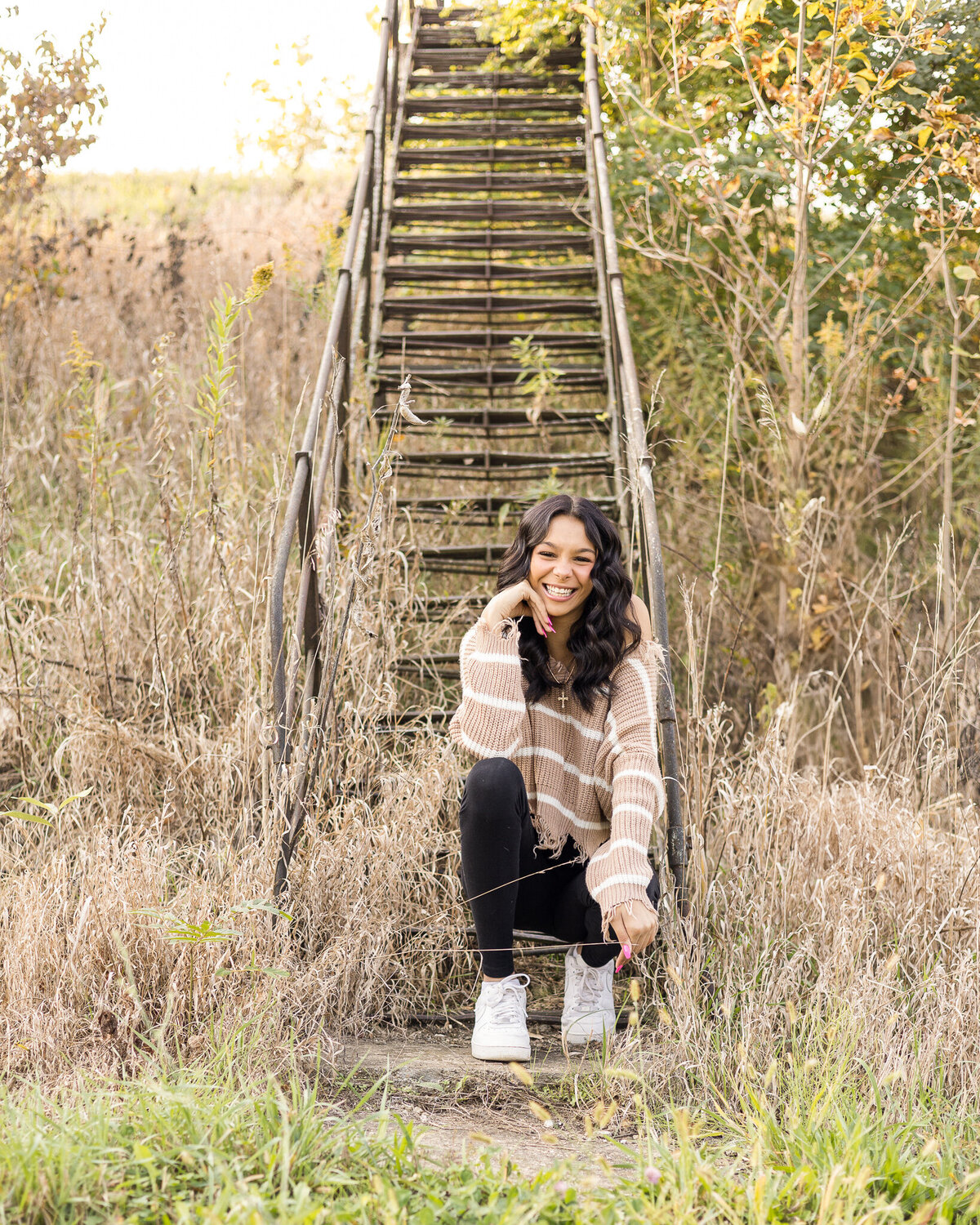 Senior female with long black hair, knelt down in front of stairs, hand under her chin, smiling at the camera, wearing black leggings, white tennis shoes, and tan and white striped long sleeve shirt