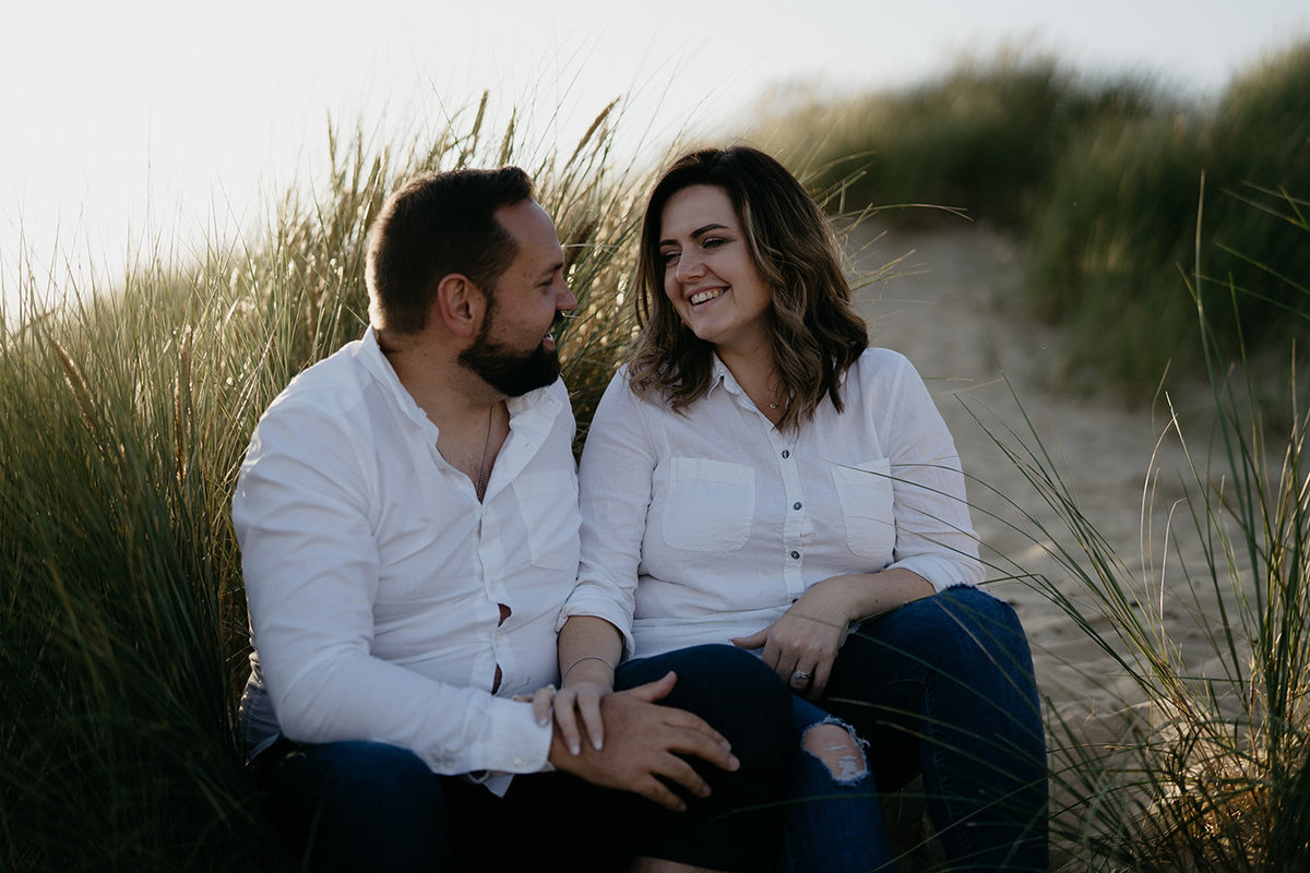 Engaged couple share a joke whilst sitting in sand dunes at Camber Sands