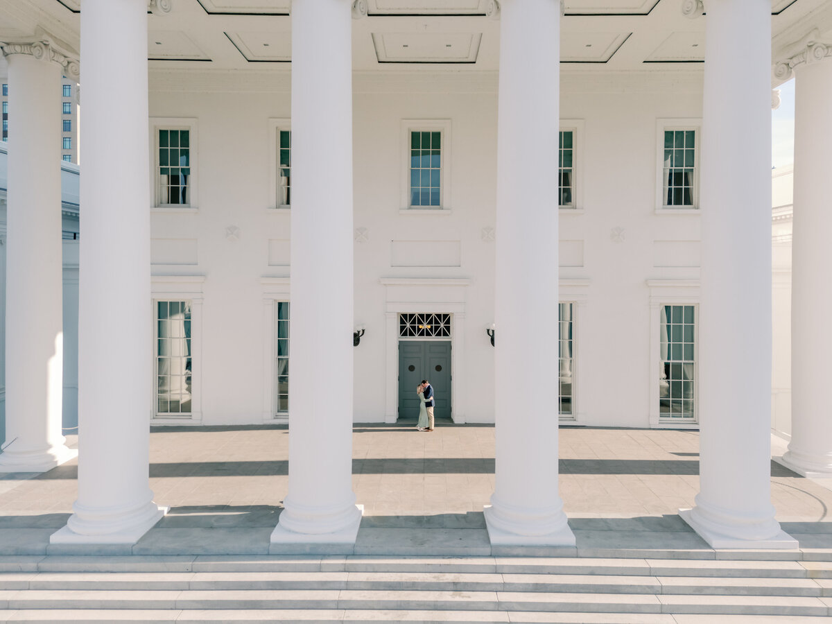 Virginia State Capitol Building Engagement Photos Richmond VA | A newly engaged couple shares a romantic moment beneath the grand white columns of the Virginia State Capitol in Richmond, Virginia. The neoclassical architecture provides an elegant and timeless backdrop, perfect for engagement photos. Captured by a Richmond engagement photographer, this image beautifully highlights one of the most historic wedding and portrait locations in Virginia.