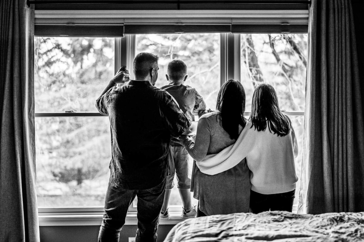A black and white photograph of a family looking out their bedroom window together during at at home photoshoot.