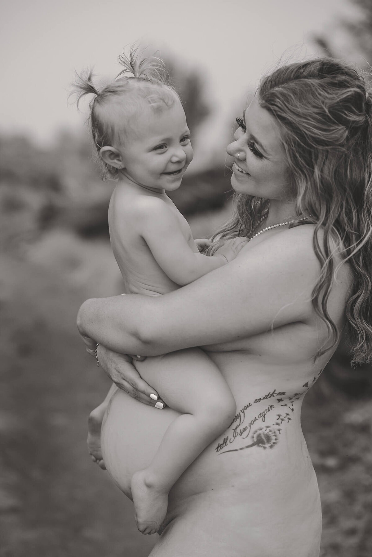 A black and white image of a nude mother and daughter. Mother is pregnant and holds her toddler on her belly while they look at eachother and smile. Taken in Tobiano, Kamloops by Brittany Danielle