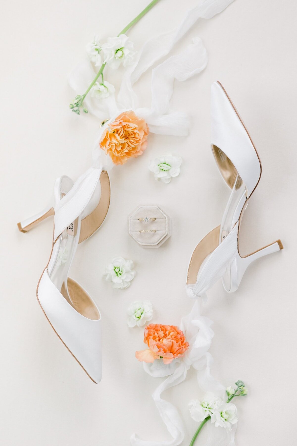 Flatlay of white satin wedding shoes with rings and flowers