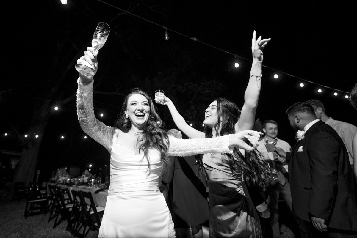 Black and white photograph of a bride and her maid of honor dancing with their hands up in the air during the wedding reception at The White Home in Rock Hill