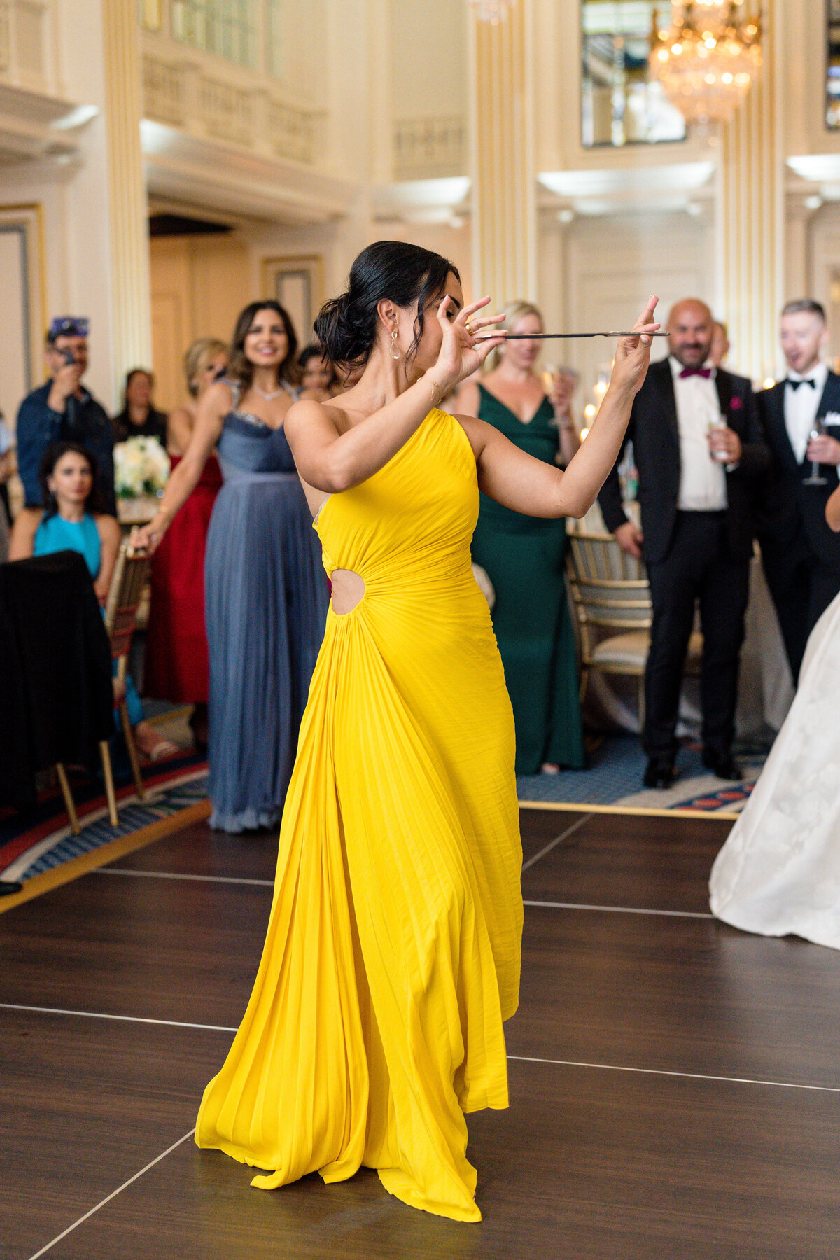 A woman in a bright yellow dress dances joyfully at an event in an elegant ballroom. Guests in formal attire watch and smile in the background, with chandeliers and luxurious decor visible.