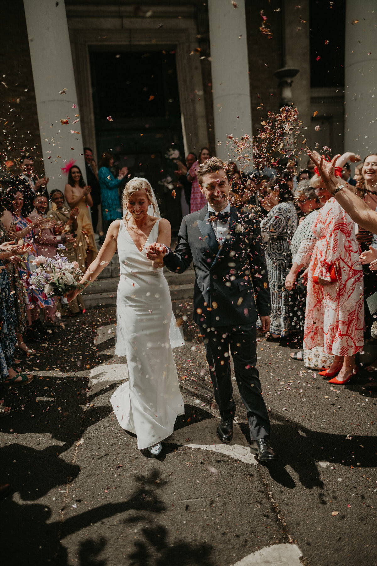 A bride and groom walk through a tunnel of their guests throwing confetti at The Asylum Chapel in London. 