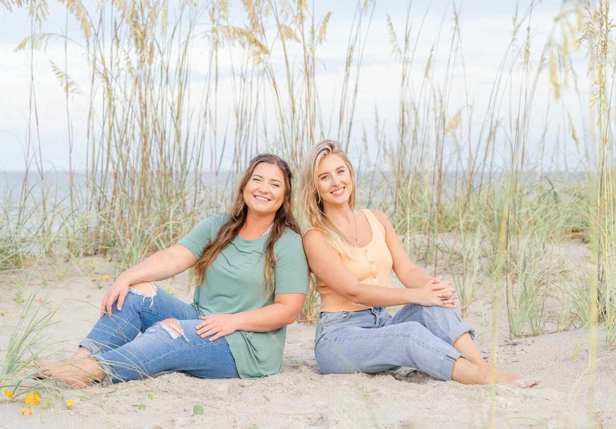 sibling sisters sitting in sand on beach with ocean in background